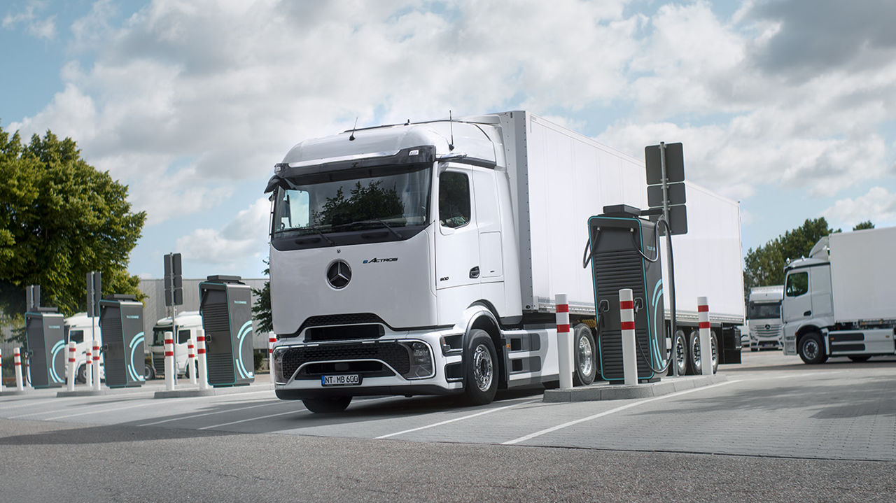 Bílý eActros 600 s ProCabin od Mercedes‑Benz Trucks s bílým návěsem stojí na parkovišti a nabíjí se na nabíjecí stanici. Kolem nákladního vozu jsou vidět další nabíjecí stanice a ochranné sloupky. V pozadí stojí další bílé nákladní vozy a na levém okraji snímku jsou pod částečně oblačnou oblohou vidět zelené stromy.