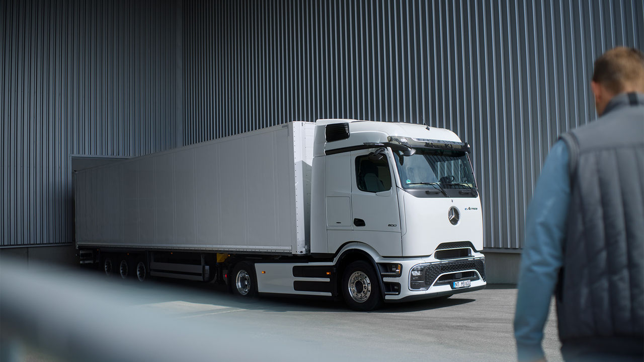 Bílý eActros 600 s ProCabin od Mercedes-Benz Trucks s dlouhým bílým návěsem stojí na asfaltované ploše před šedou vlnitou plechovou stěnou. Kamion je vidět ze strany, přičemž jeho příď je mírně natočená doleva. V popředí, se zády k pozorovateli, stojí osoba v modré bundě a šedé vestě, která se dívá na kamion. Světlo dopadá shora na scénu.