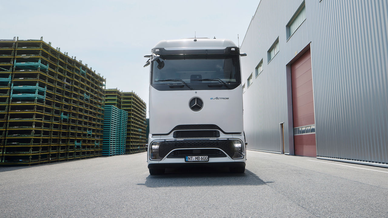 eActros 600 s ProCabin od Mercedes‑Benz Trucks stojí čelně na asfaltovaném prostranství. Vlevo v záběru jsou vidět vysoké stohy žlutých a modrých palet, zatímco vpravo stojí velká moderní budova s šedou fasádou z vlnitého plechu a červenými vraty. Obloha je světlá a lehce oblačná.