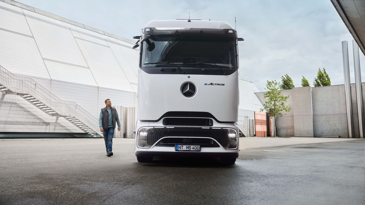 Muž v pracovním oděvu jde po asfaltované ploše, zatímco v pozadí parkuje bílý eActros 400 s ProCabin od Mercedes-Benz Trucks. Vlevo je vidět moderní budova s dlouhým venkovním schodištěm a vpravo stromy a další stavební objekty.