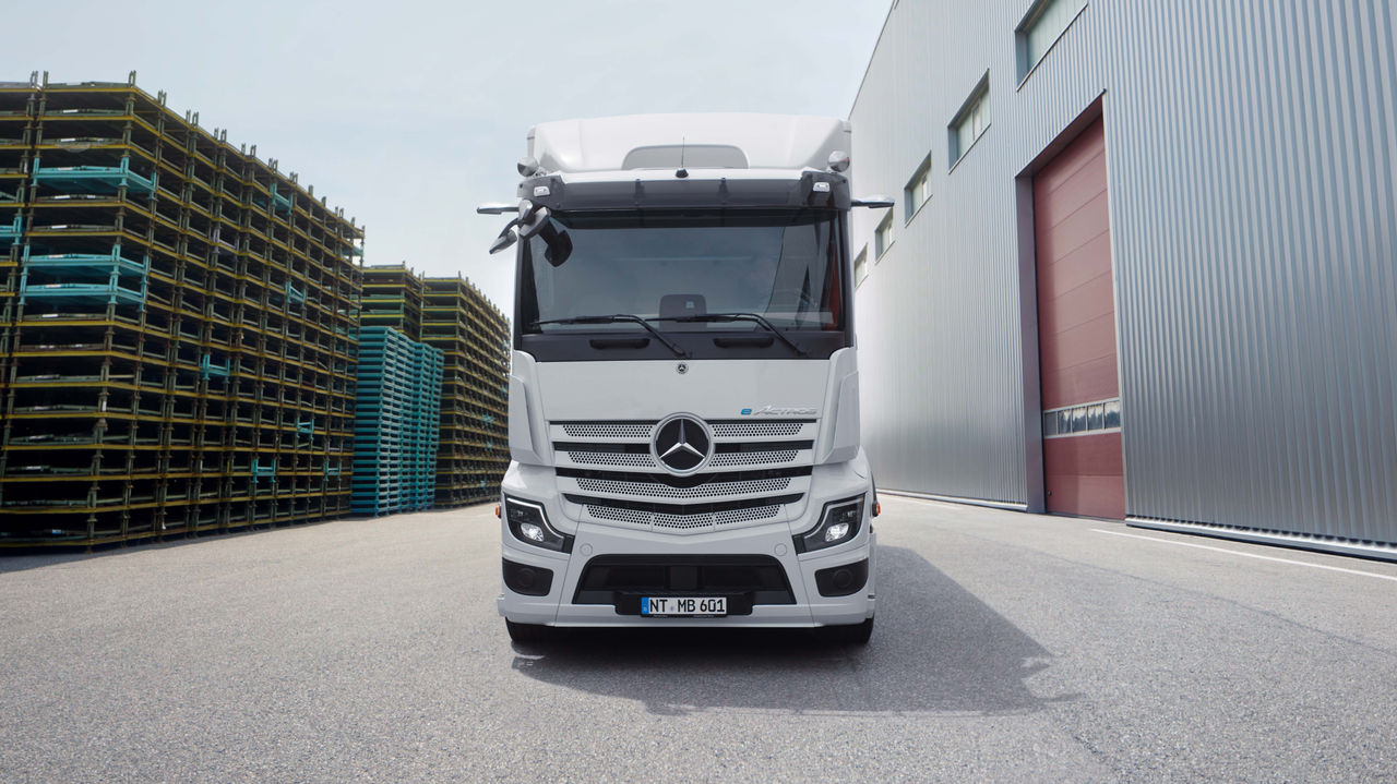 Před moderní budovou s dlouhým vnějším schodištěm čelně stojí na asfaltové ploše eActros 600 od Mercedes-Benz Trucks. Nákladní vůz je bílý a má na mřížce chladiče logo hvězdy Mercedesu, zatímco v pozadí jsou vidět stromy a další budovy.