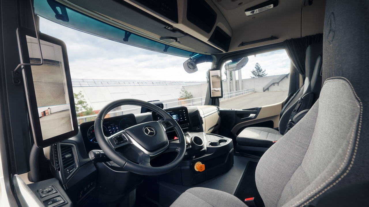 Snímek zobrazuje interiér eActros od Mercedes-Benz Trucks z pohledu sedadla spolujezdce. Je vidět volant s hvězdou Mercedesu, přístrojová deska s digitálními ukazateli a displeji a také pohodlná sedadla z světle šedé tkaniny.