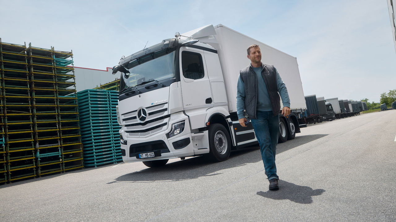 Muž v pracovním oblečení kráčí po asfaltované ploše, zatímco v pozadí parkuje bílý eActros 600 od Mercedes-Benz Trucks s přívěsem. Vlevo jsou naskládané palety a vpravo stojí velká budova, která naznačuje skladový nebo průmyslový areál.