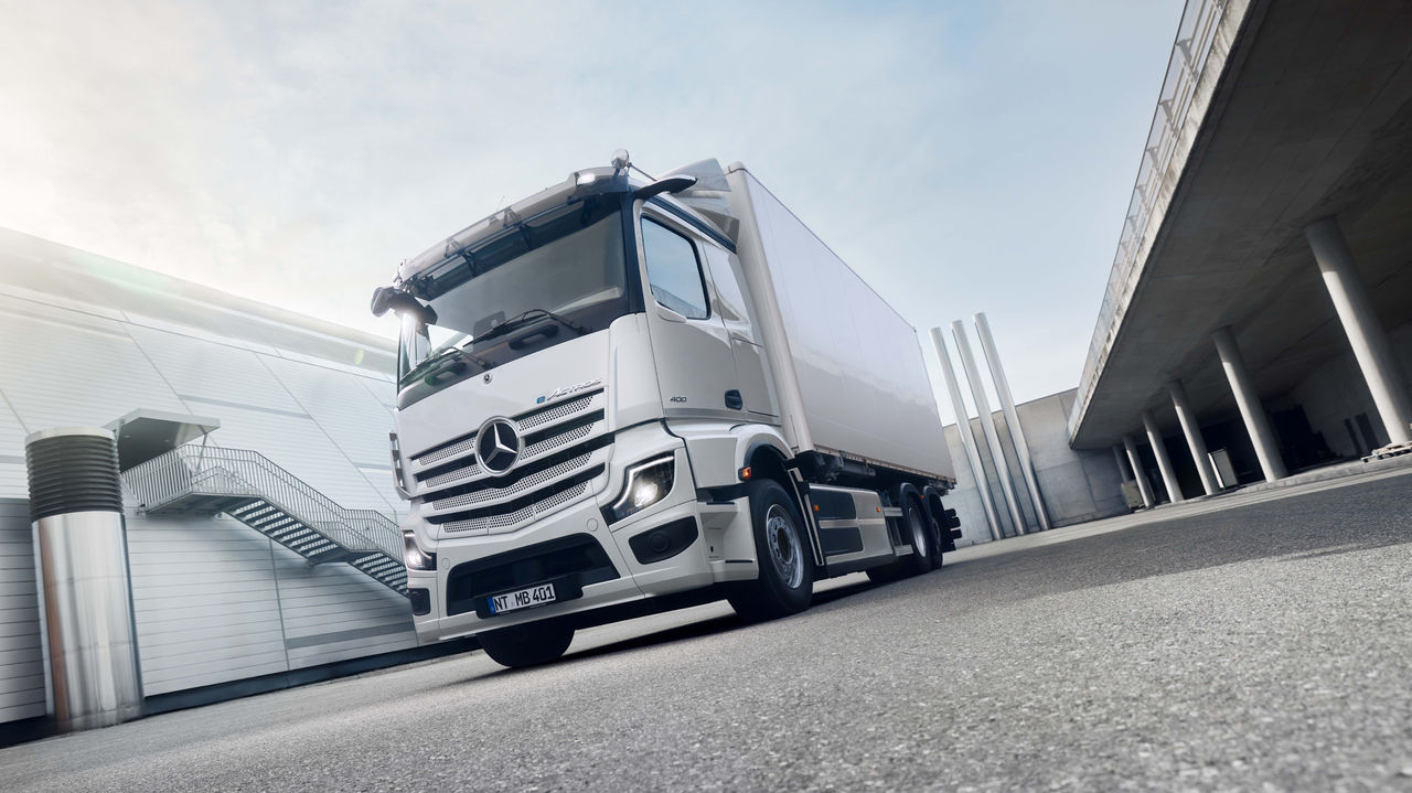 Bílý eActros 400 od Mercedes‑Benz Trucks stojí na asfaltované ploše před moderní budovou s venkovním schodištěm, přičemž slunce jasně svítí zleva shora. Vpravo v záběru je vidět zvýšená betonová konstrukce s pilíři, která naznačuje podchod nebo zastřešenou plochu.