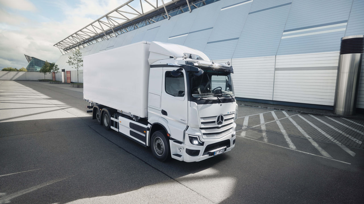 Bílý eActros 400 od Mercedes‑Benz Trucks se skříňovou nástavbou stojí na asfaltovaném parkovišti před moderní budovou se skleněnou fasádou. Sluneční světlo vrhá dlouhé stíny na zem, což naznačuje jasný den.